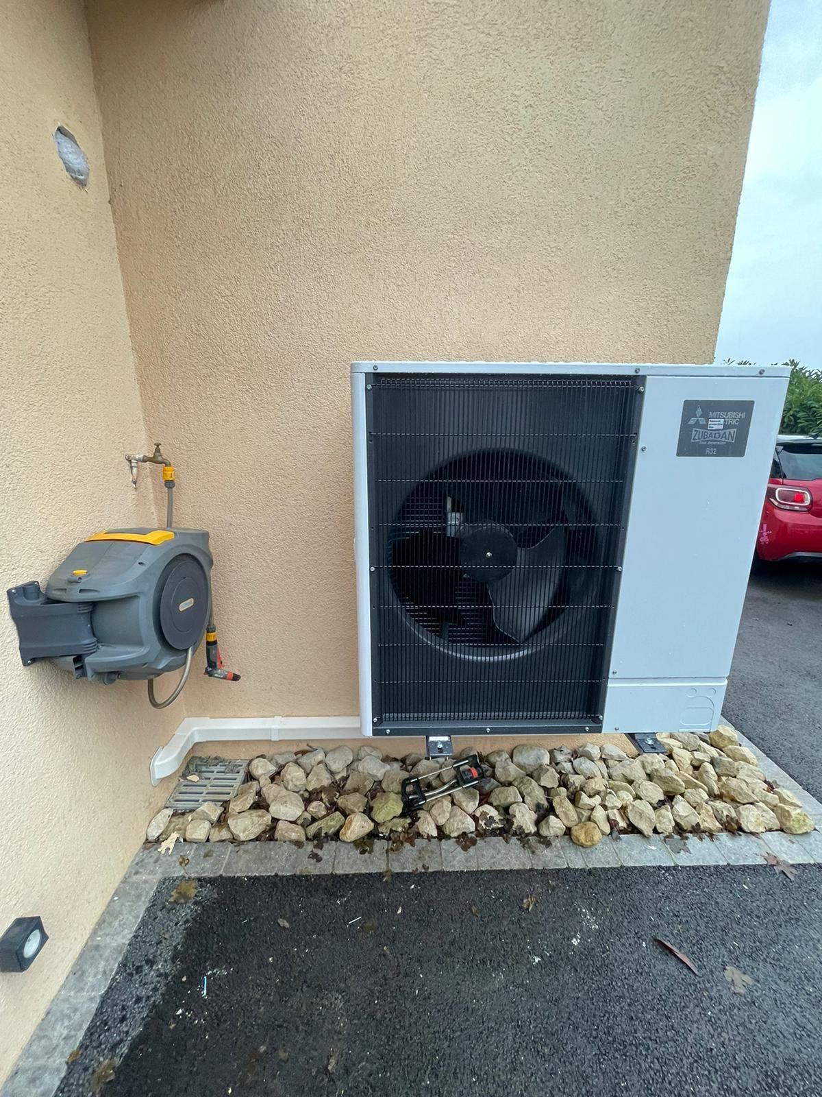 Unité de climatisation extérieure à côté d'un mur en stuc beige, avec un compteur de gaz et un lit de pierres le long du bâtiment