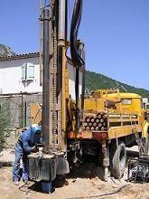 Une foreuse jaune avec un ouvrier en salopette bleue, devant un bâtiment blanc par une journée ensoleillée.