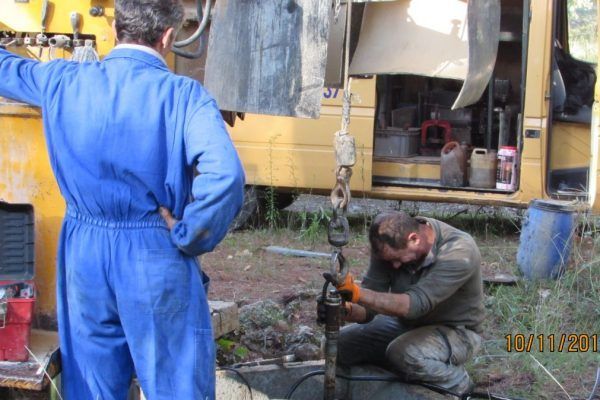 Deux ouvriers près d'une foreuse. L'un est agenouillé et travaille. L'autre, debout, observe. En extérieur.