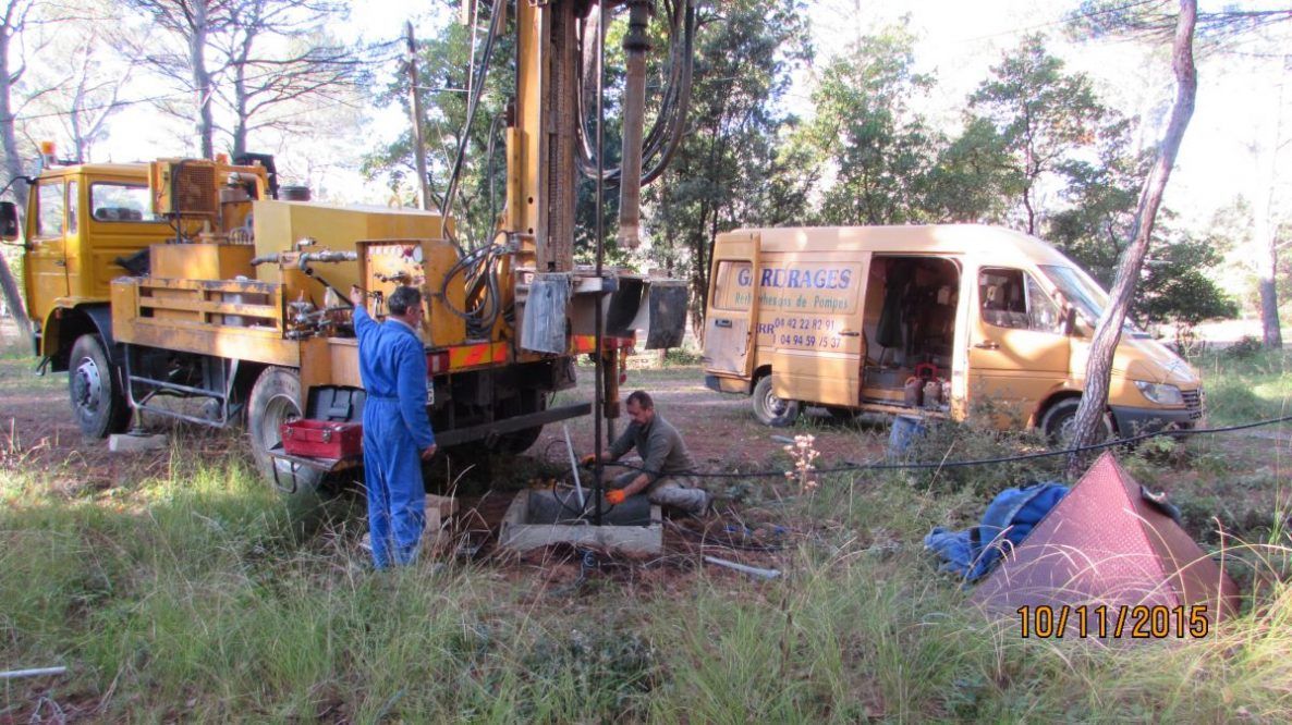 Des ouvriers effectuent des forages avec des engins dans une zone boisée ; un camion et une camionnette sont stationnés à proximité.