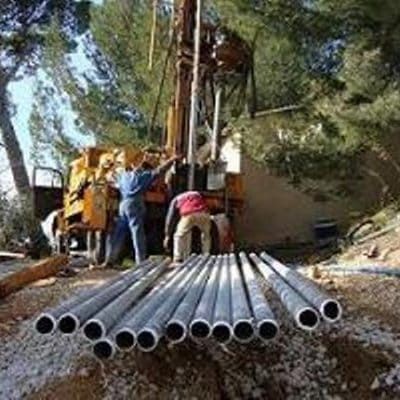 Des ouvriers forent un puits à l'aide d'une foreuse. Des tuyaux sont posés au sol. Des arbres se dressent en arrière-plan.