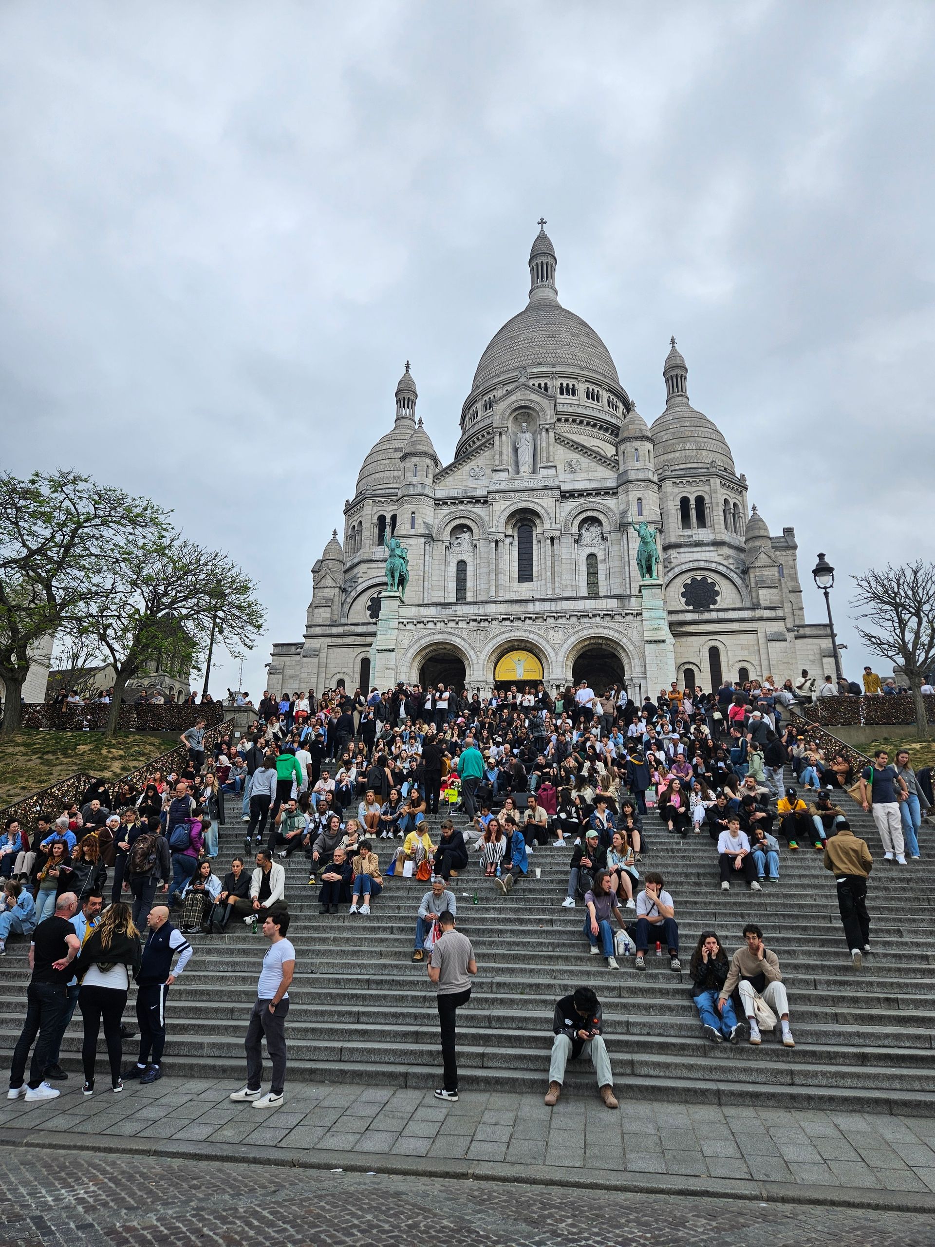 Foule massée sur de larges marches devant le Sacré-Cœur, sous un ciel nuageux.