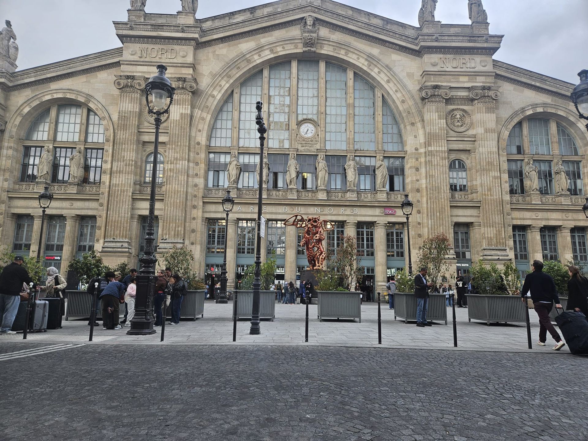 Façade historique en pierre de la gare du Nord, avec de grandes fenêtres cintrées et des personnes rassemblées sur une place à l'extérieur.