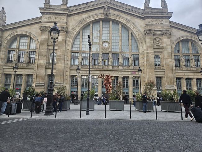Façade historique en pierre de la gare du Nord, avec de grandes fenêtres cintrées et des personnes rassemblées sur une place à l'extérieur.