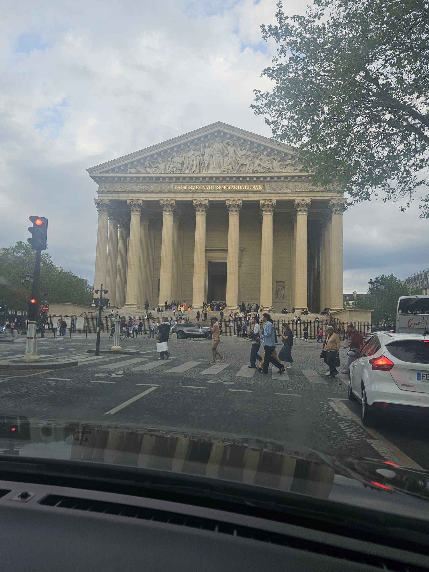 Foule traversant devant l'église de la Madeleine à Paris, vue depuis une voiture dans un trafic pluvieux.