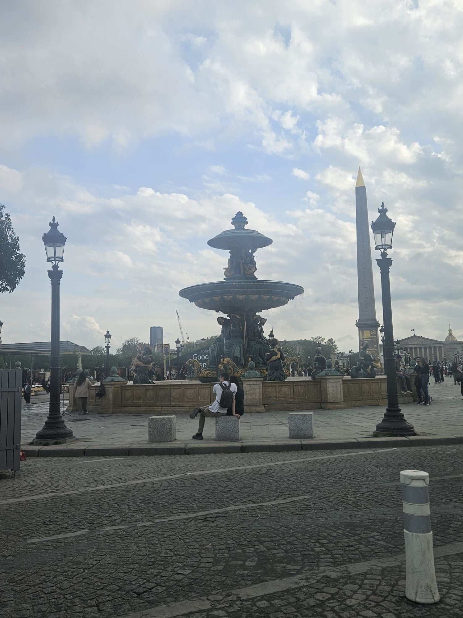 Fontaine de la Concorde sous un ciel nuageux, avec des passants à proximité et des lampadaires ornementés autour.