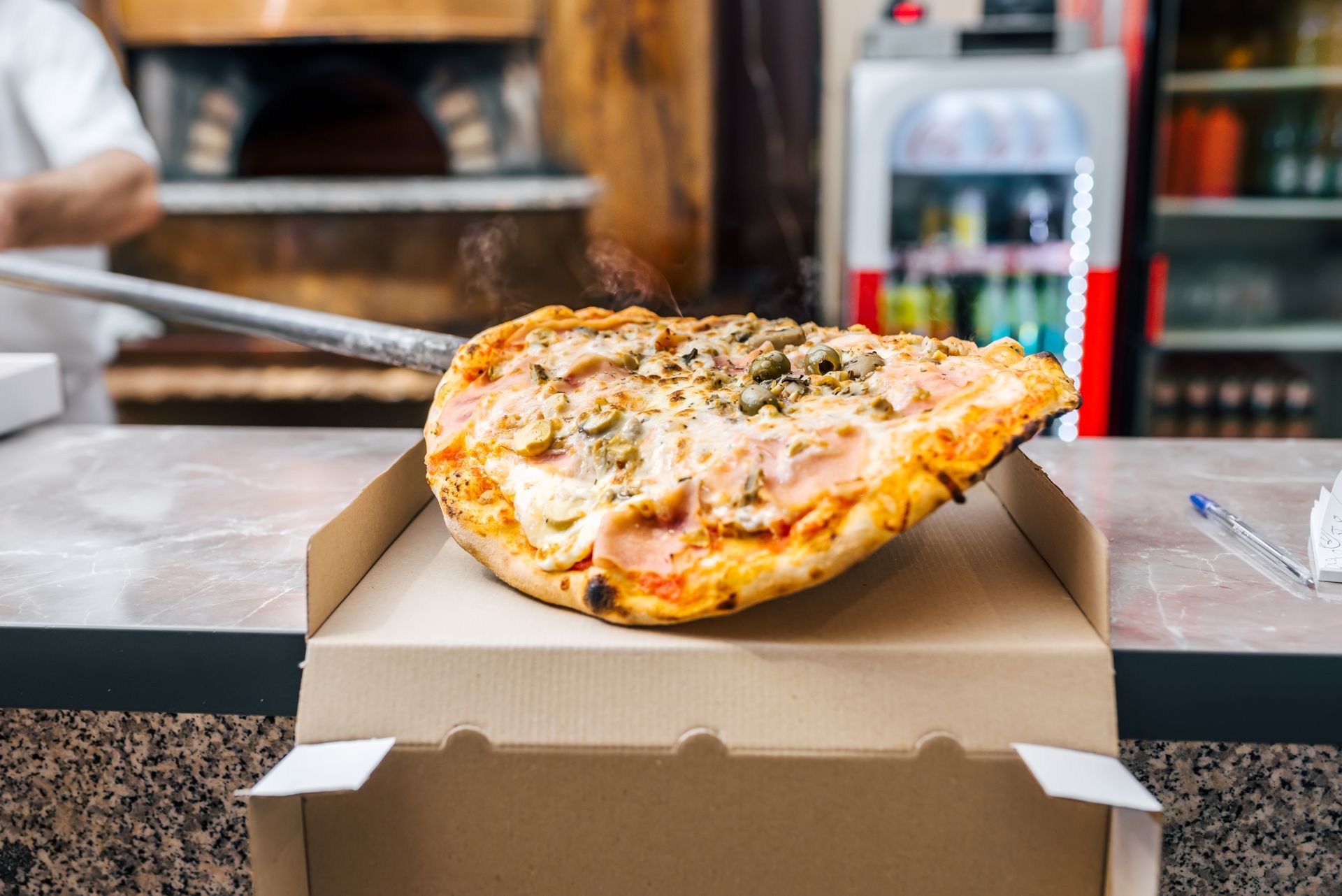 Pizza fraîchement cuite sur une boîte en carton, maintenue par une pelle. De la fumée s'élève.
