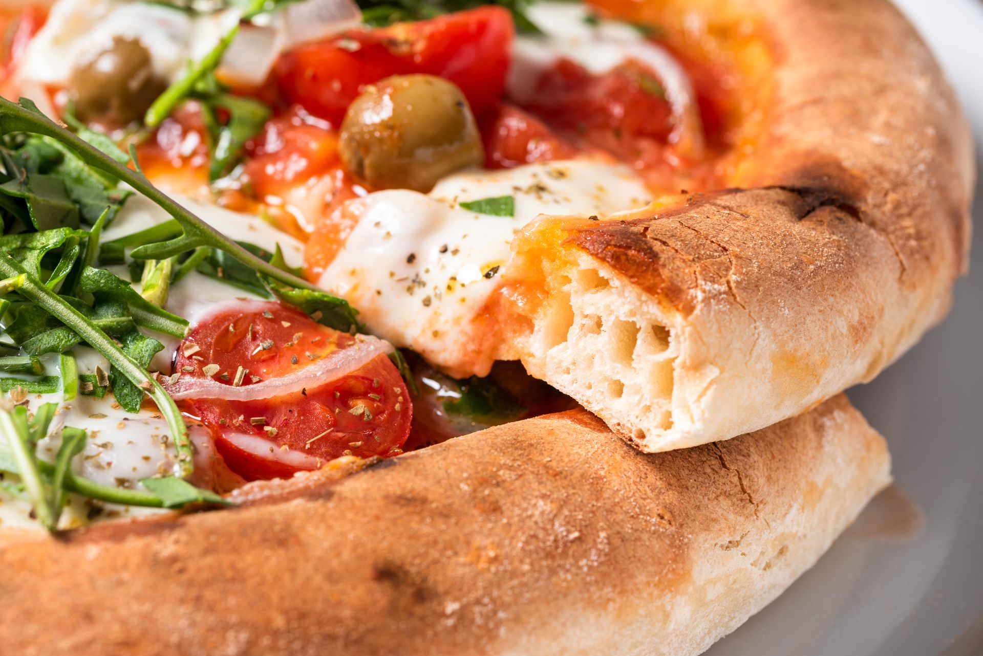 Gros plan d'une pizza avec des tomates, des olives, de la roquette et du fromage ; la croûte est déchirée pour révéler des poches d'air.