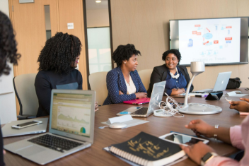 A diverse group of women in business meeting at a conference table with laptops and a whiteboard.