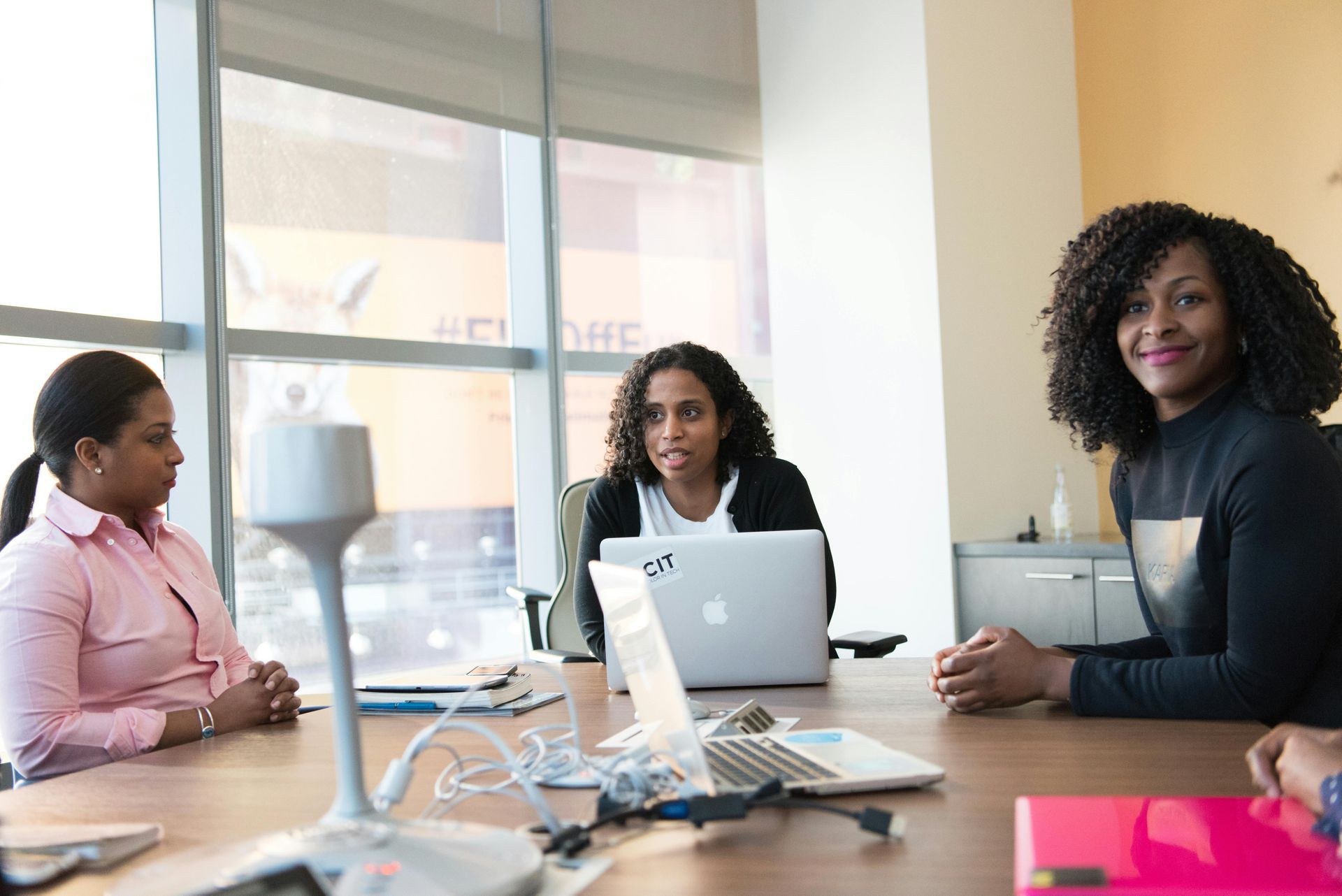 A group of women are sitting at a conference table with laptops.