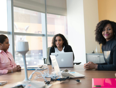 Three women in a meeting room, two looking at laptop, one smiling, all are women of color, sunlight streaming through windows