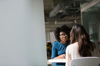 Two women at a table in an office. One looks towards the other, wearing glasses and a blue shirt.