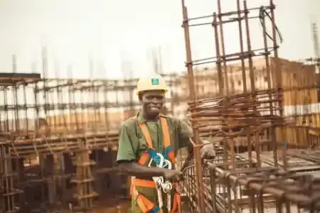 Construction worker in yellow hard hat and safety harness smiles at the camera on a construction site.