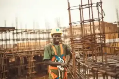 Construction worker in yellow hard hat and safety harness smiles at the camera on a construction site.