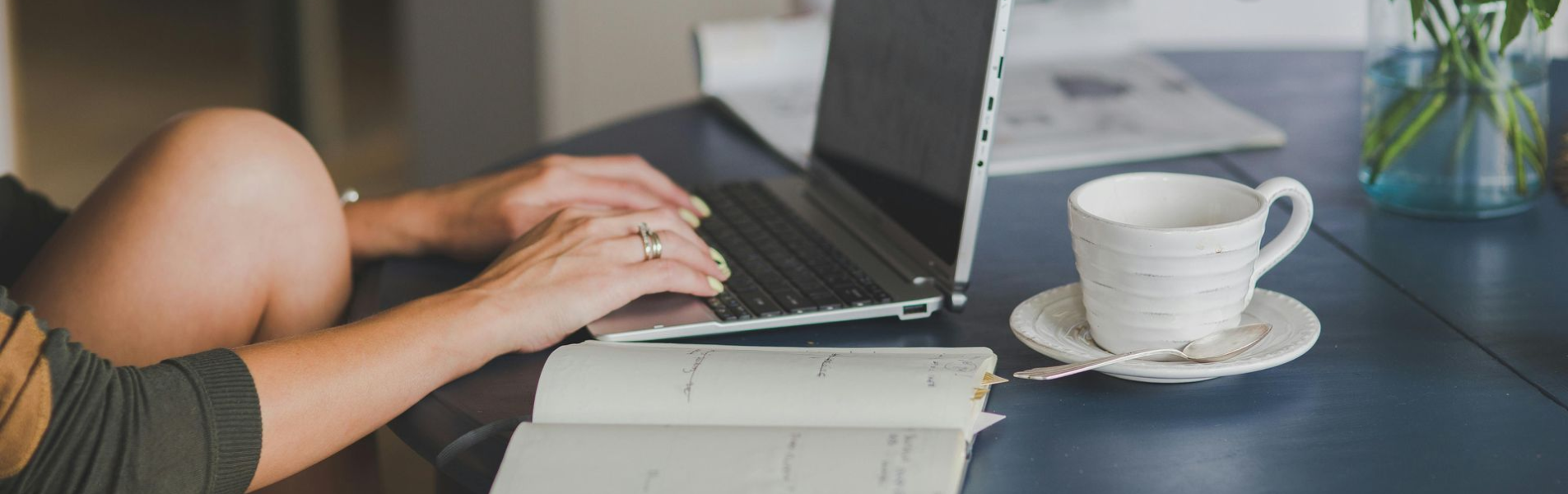 Woman typing on a laptop at a blue table, with coffee, flowers, and a notebook in a home setting.