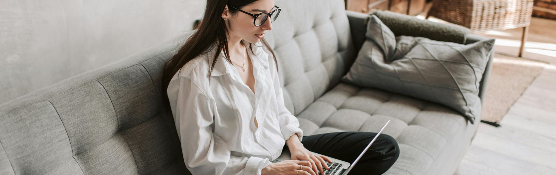Woman with glasses works on laptop, sitting on a gray sofa in a home setting.