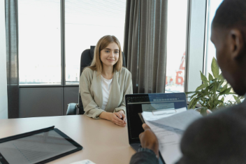 Woman in a light blazer smiles during a job interview, seated at a desk across from an interviewer holding papers.
