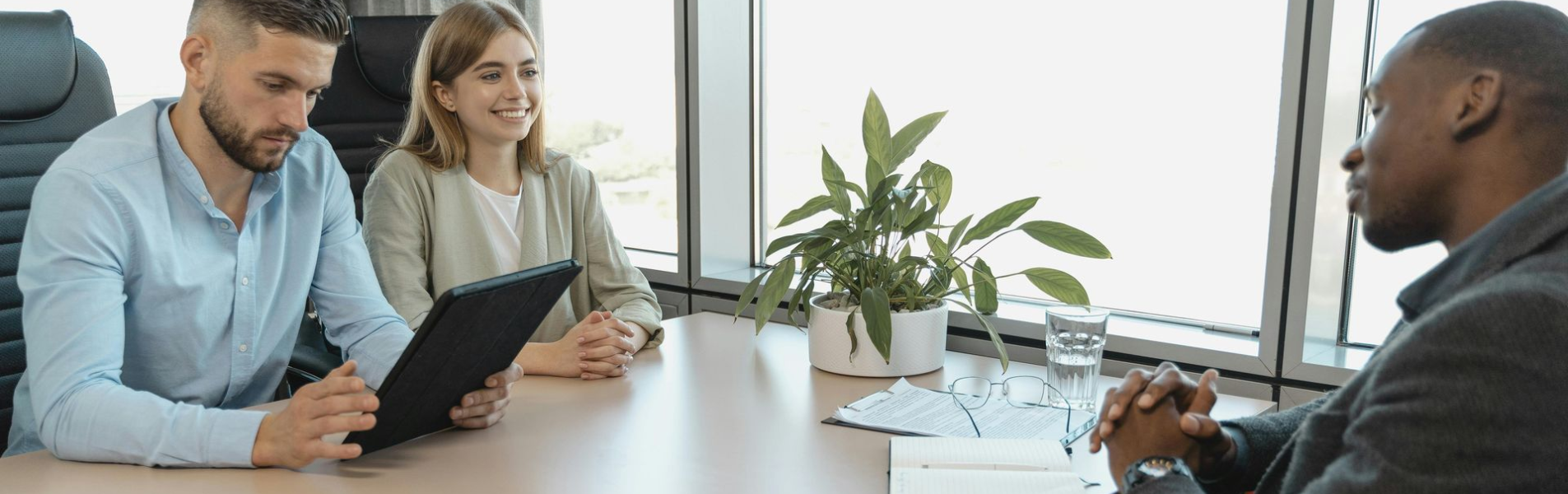 Three people in an office setting. Two interviewers with a tablet face a person with hands clasped, by a window.