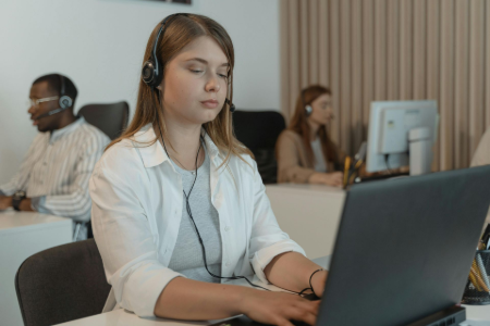 Woman with curly hair wearing a headset smiles, working at a desk. She wears a yellow shirt.