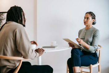 Woman interviewing a man at a table. Woman holds clipboard and pen. White walls, neutral colors.