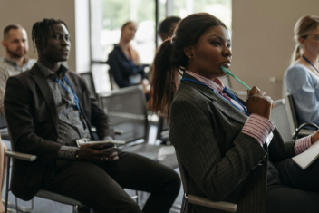 Attendees at a conference, the woman in the foreground holds a pen to her mouth, the man next to her looks forward.
