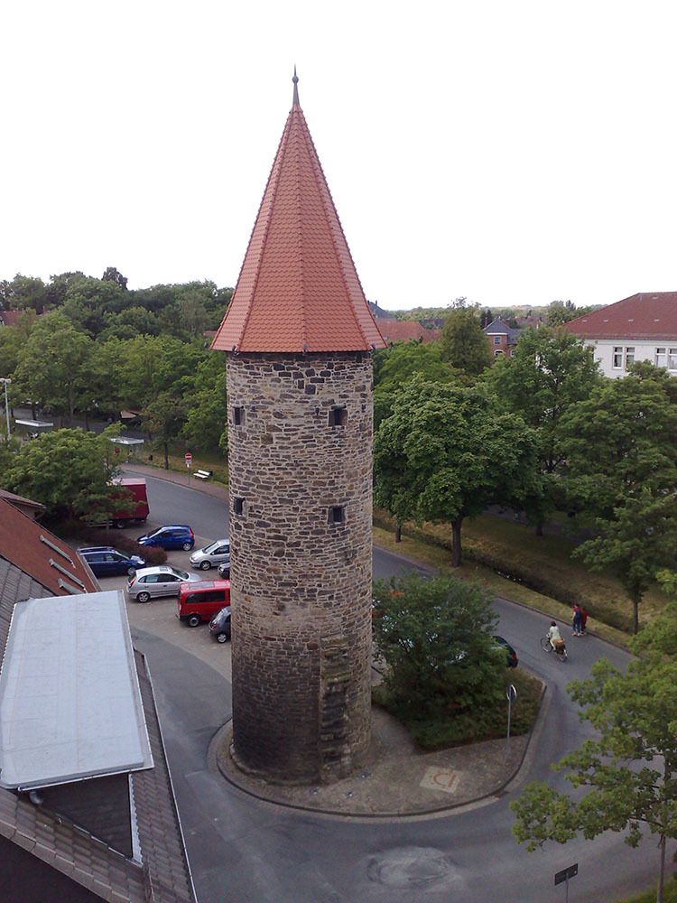 Steinturm mit rotem Dach auf einem Stadtplatz, umgeben von Autos und Bäumen.