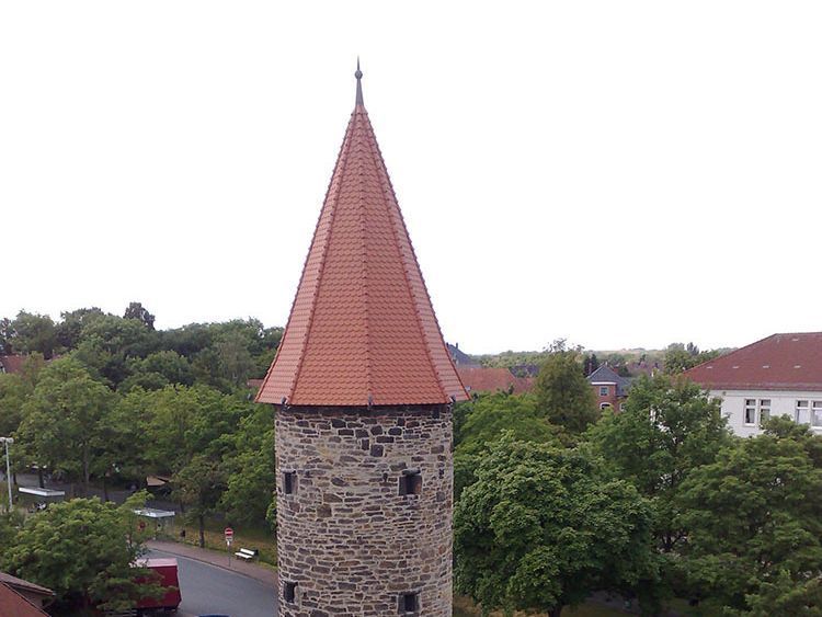 Steinturm mit rotem Ziegeldach, Bäumen und bewölktem Himmel.