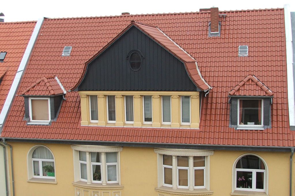 Gelbes Gebäude mit rotem Ziegeldach, Dachgauben und schwarzen Fassadenakzenten.