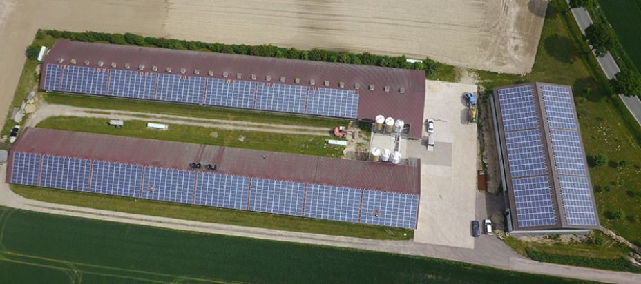Luftaufnahme von landwirtschaftlichen Gebäuden mit Solarmodulen. Die Gebäude sind rechteckig mit braunen Dächern und liegen neben Feldern.