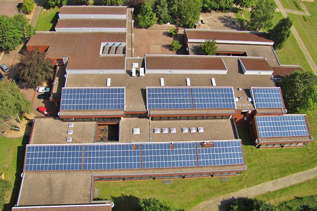 Luftaufnahme eines Schulgebäudes mit Solarmodulen auf dem Dach, umgeben von grünem Gras und Bäumen.