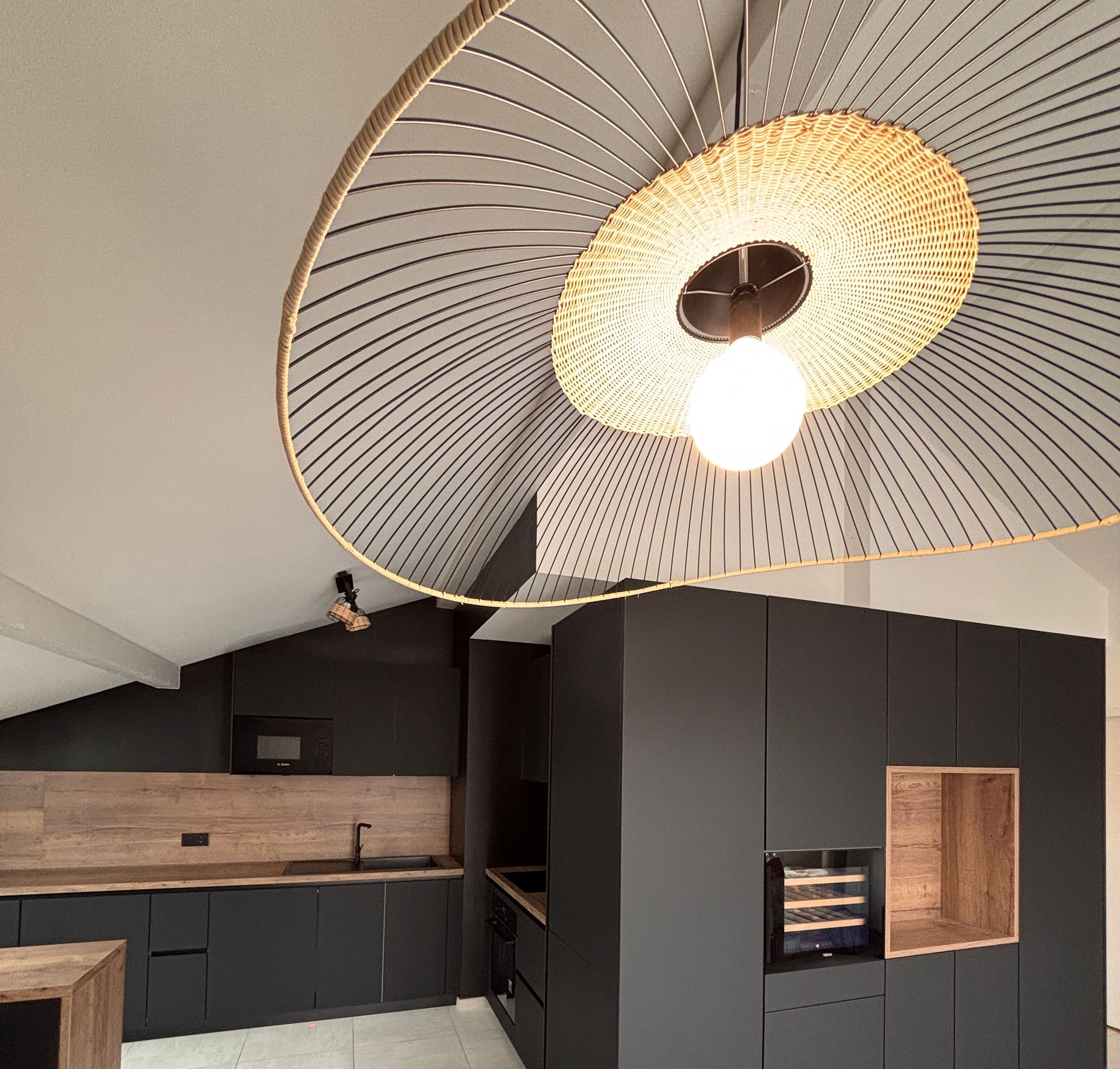 Cuisine moderne avec placards foncés, plan de travail et étagères ouvertes, grand luminaire suspendu en tissu tressé.