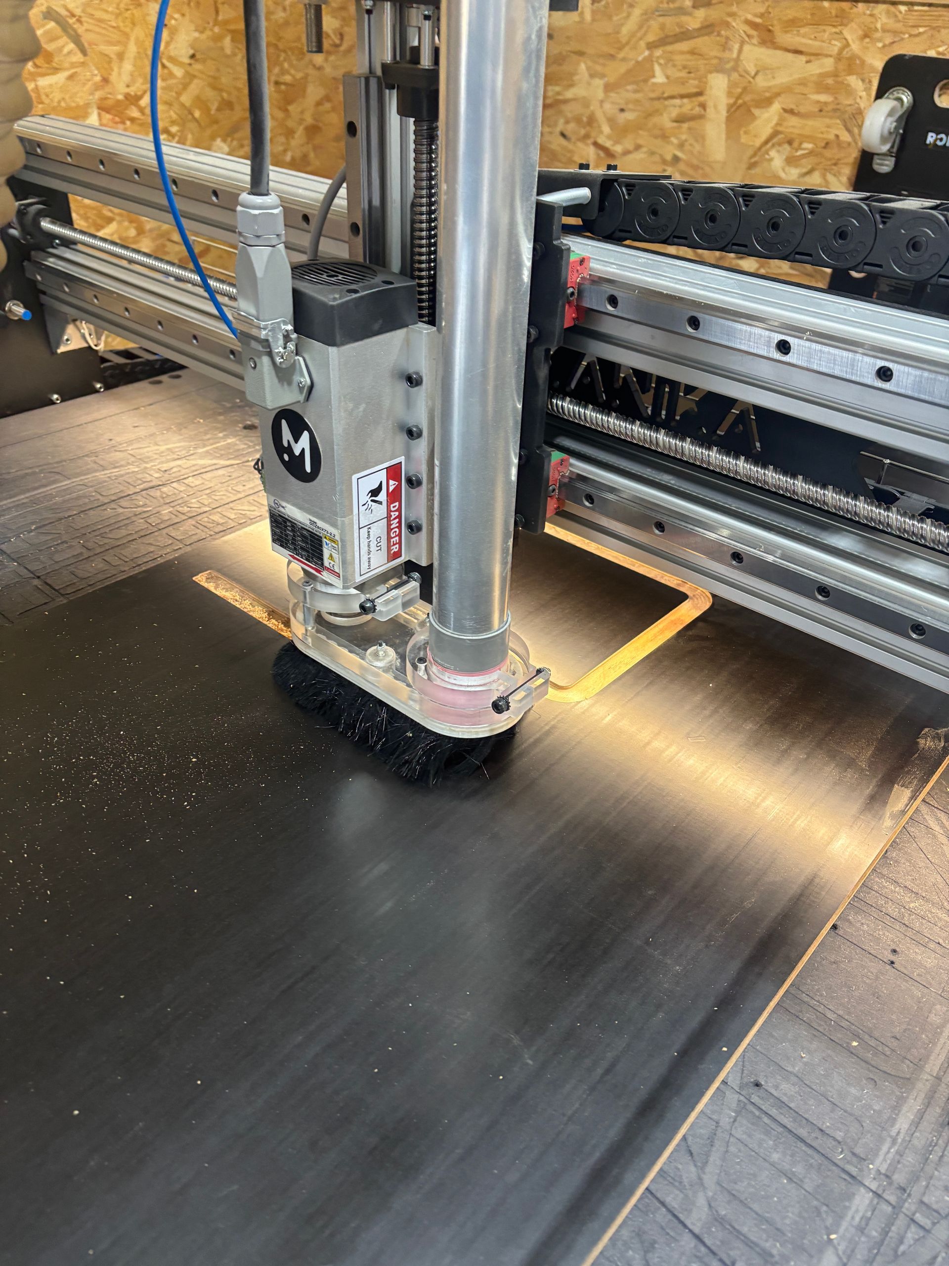 Une fraiseuse CNC découpe un carré dans du bois. Une brosse entoure la fraise en rotation. De la sciure est visible.