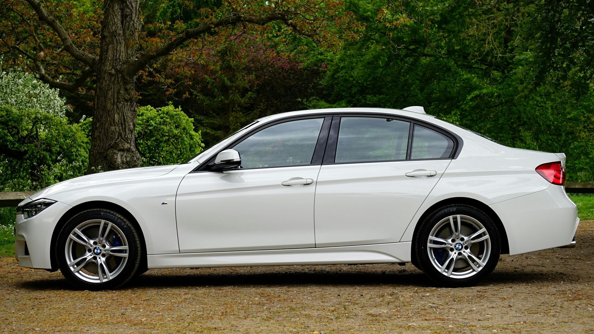 Eine weiße BMW-Limousine parkt auf einem Schotterweg; im Hintergrund sind Bäume zu sehen.