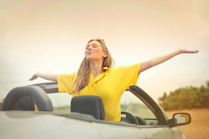 Frau im gelben Hemd, die Arme ausgestreckt, in einem Cabriolet, sonnenbeschienener Himmel.