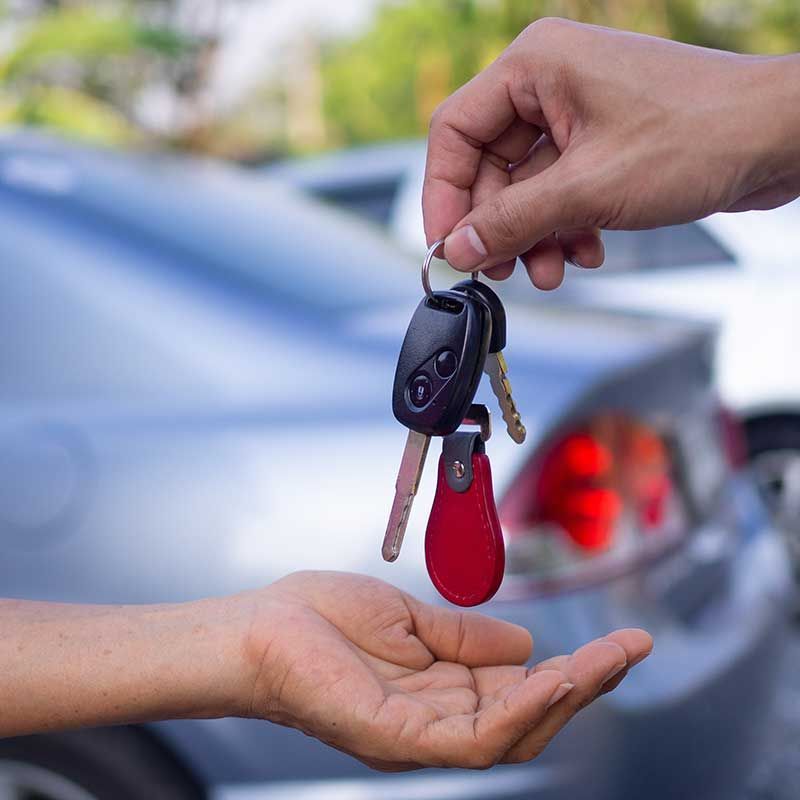 Die Hand einer Person übergibt einer anderen Person die Autoschlüssel. Im Hintergrund ist das Auto zu sehen.