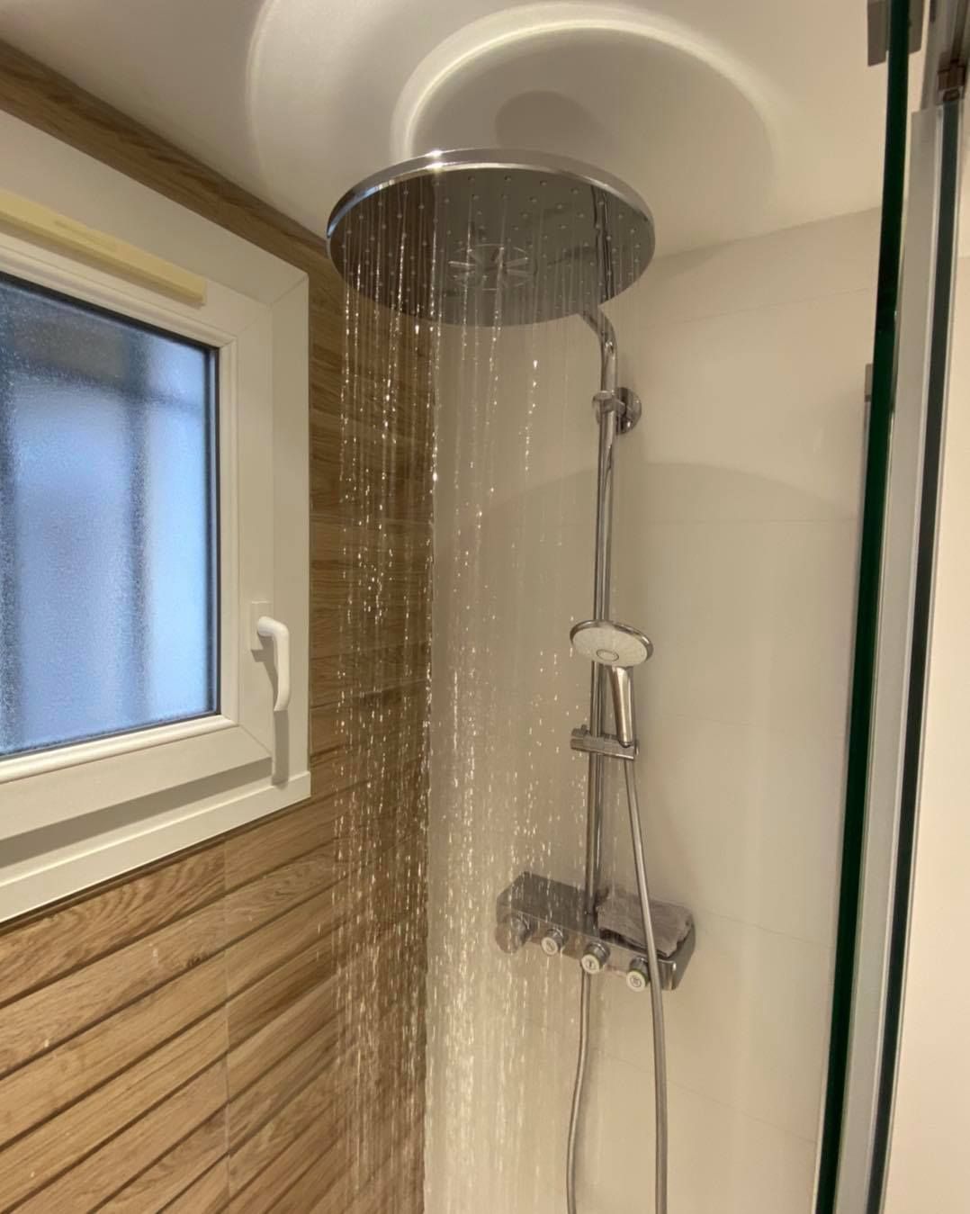 Une pomme de douche ronde à effet pluie, fixée au mur par des lattes de bois, l'eau s'écoulant dans la cabine de douche.