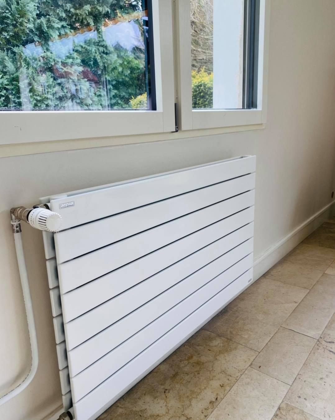 Un radiateur blanc, horizontal, à panneaux, avec un bouton de thermostat, fixé sur un mur clair sous une fenêtre.