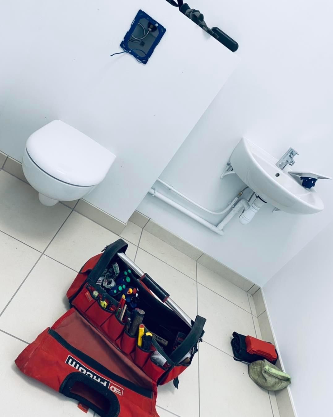 Dans une salle de bains, une sacoche à outils ouverte est posée sur le sol devant des toilettes et un lavabo fixés au mur.