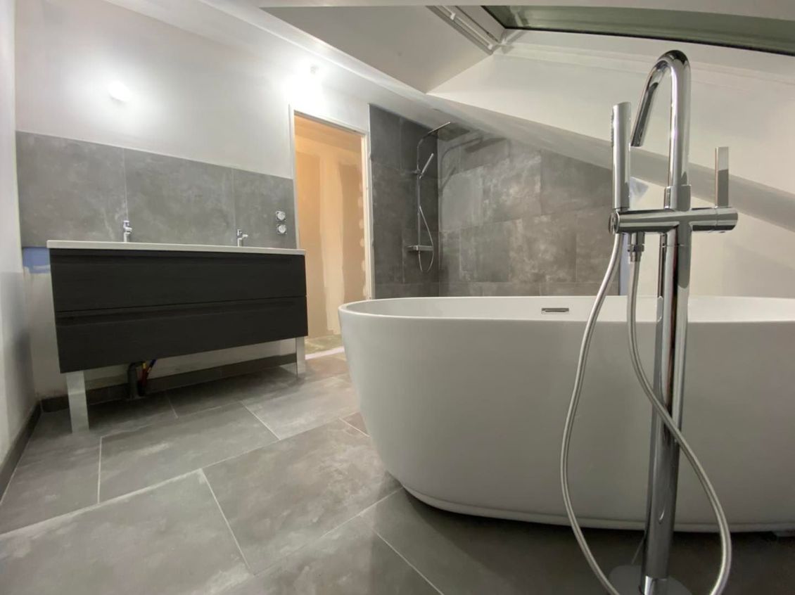 Salle de bains avec baignoire à îlot blanche et robinet chromé, meuble-lavabo gris et carrelage au sol imitation pierre.