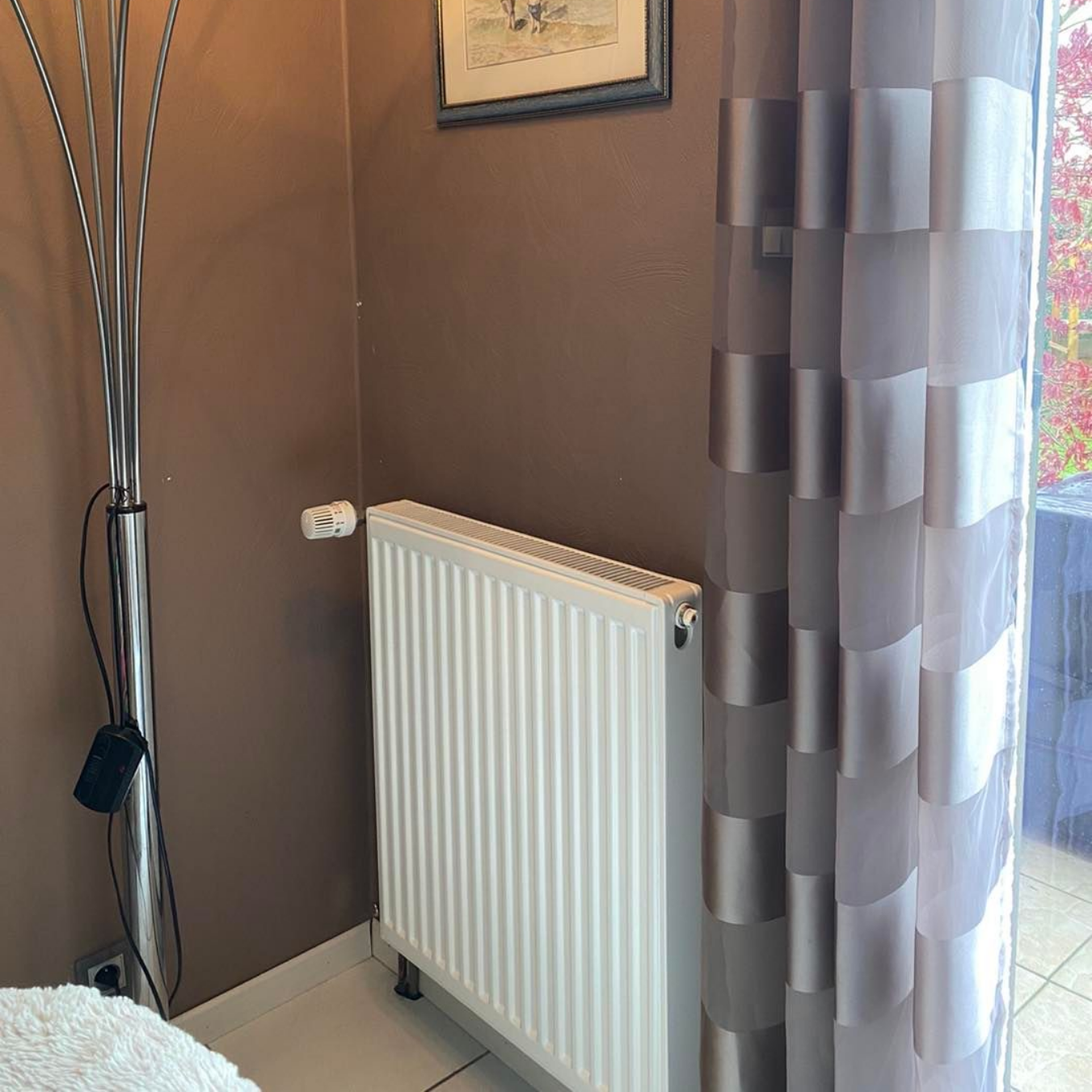 Un radiateur blanc sur un mur marron, avec un rideau transparent à rayures verticales à côté.