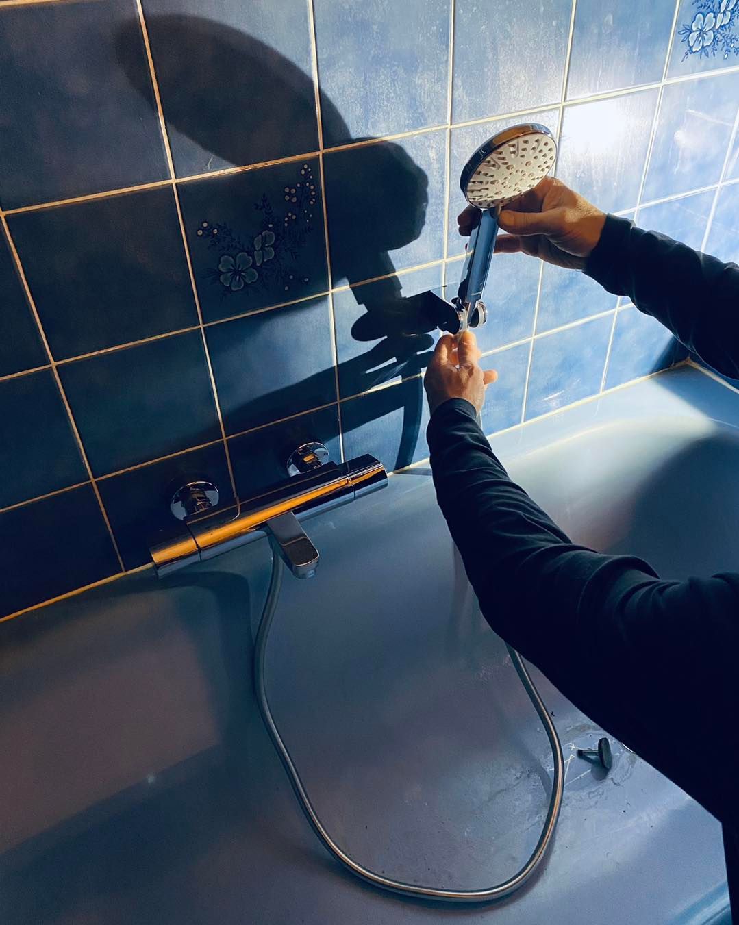 Des mains serrent un pommeau de douche argenté sur un tuyau fixé à un robinet mural, devant des murs carrelés bleu foncé.