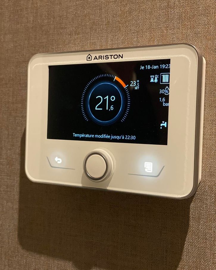 Un thermostat numérique Ariston, fixé sur un mur texturé, affiche une température de 21 degrés.