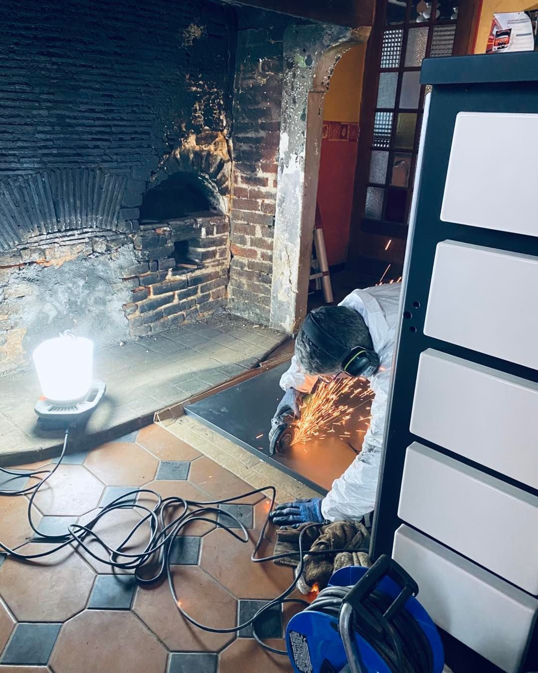 Dans une pièce rustique avec une cheminée en briques, une personne utilise une meuleuse pour découper du métal