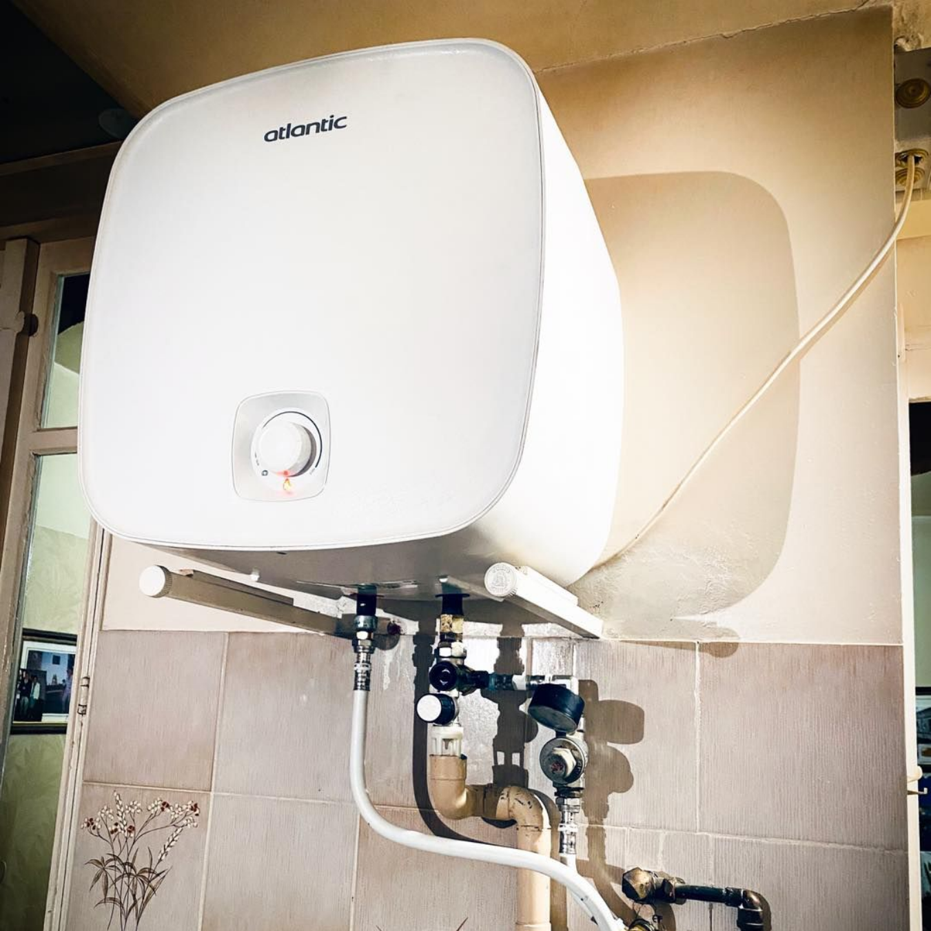 Un chauffe-eau électrique Atlantic monté sur un mur carrelé, raccordé à des tuyaux et des vannes situés en dessous.