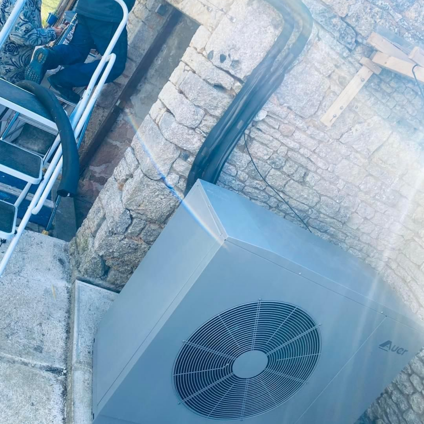 Une unité de pompe à chaleur extérieure grise est adossée à un mur de pierre, à côté d'un escalier.