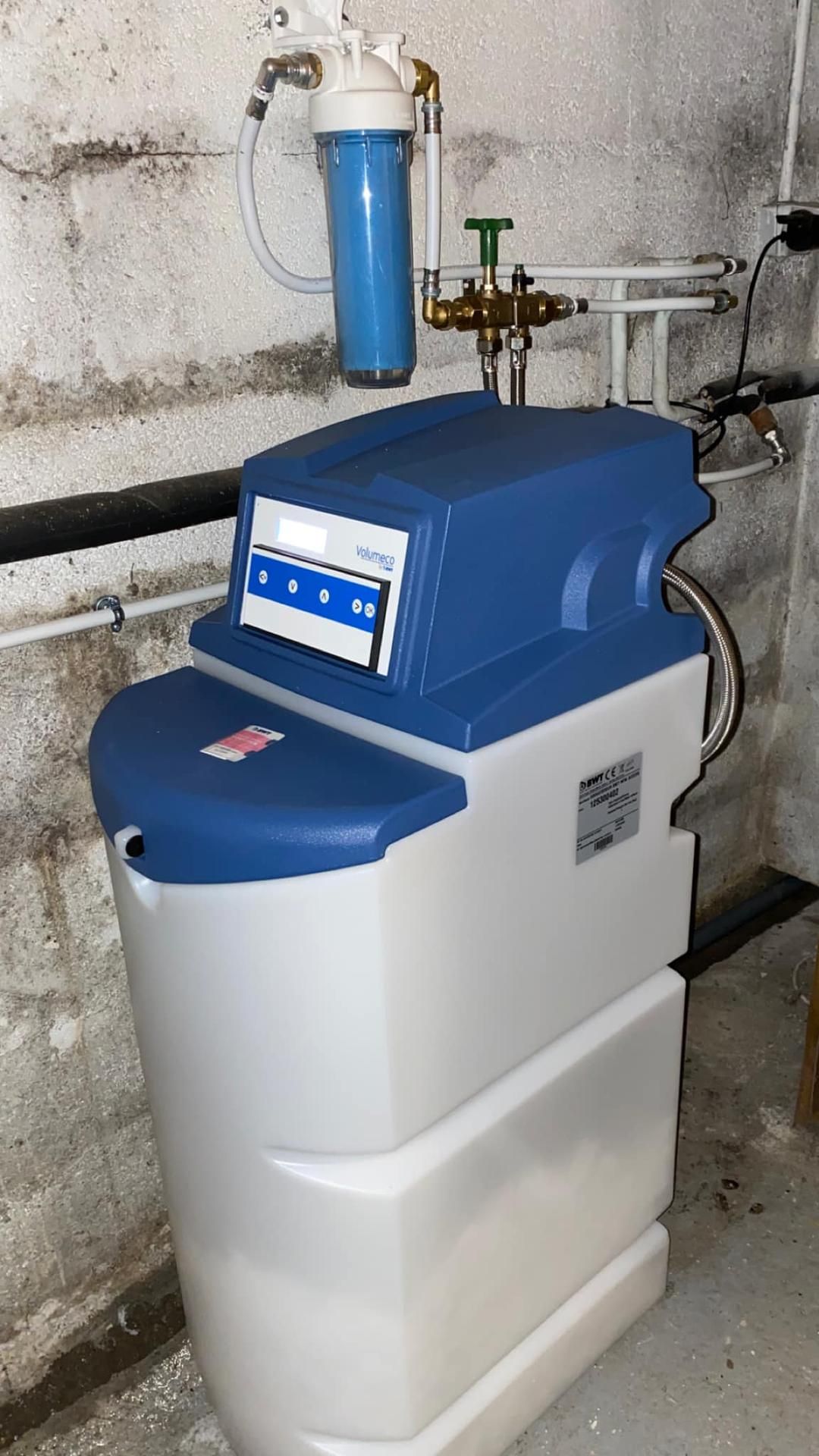 Un adoucisseur d'eau bleu et blanc avec un filtre à sédiments bleu intégré, installé contre un mur en parpaings.