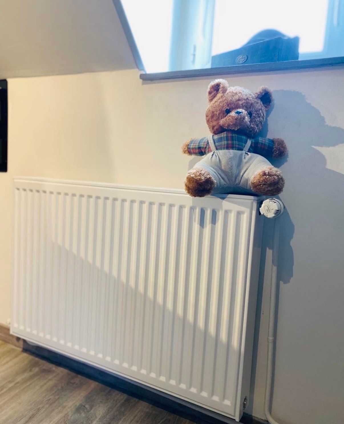 Un radiateur blanc, horizontal, à panneaux, avec un bouton de thermostat, fixé sur un mur clair sous une fenêtre.
