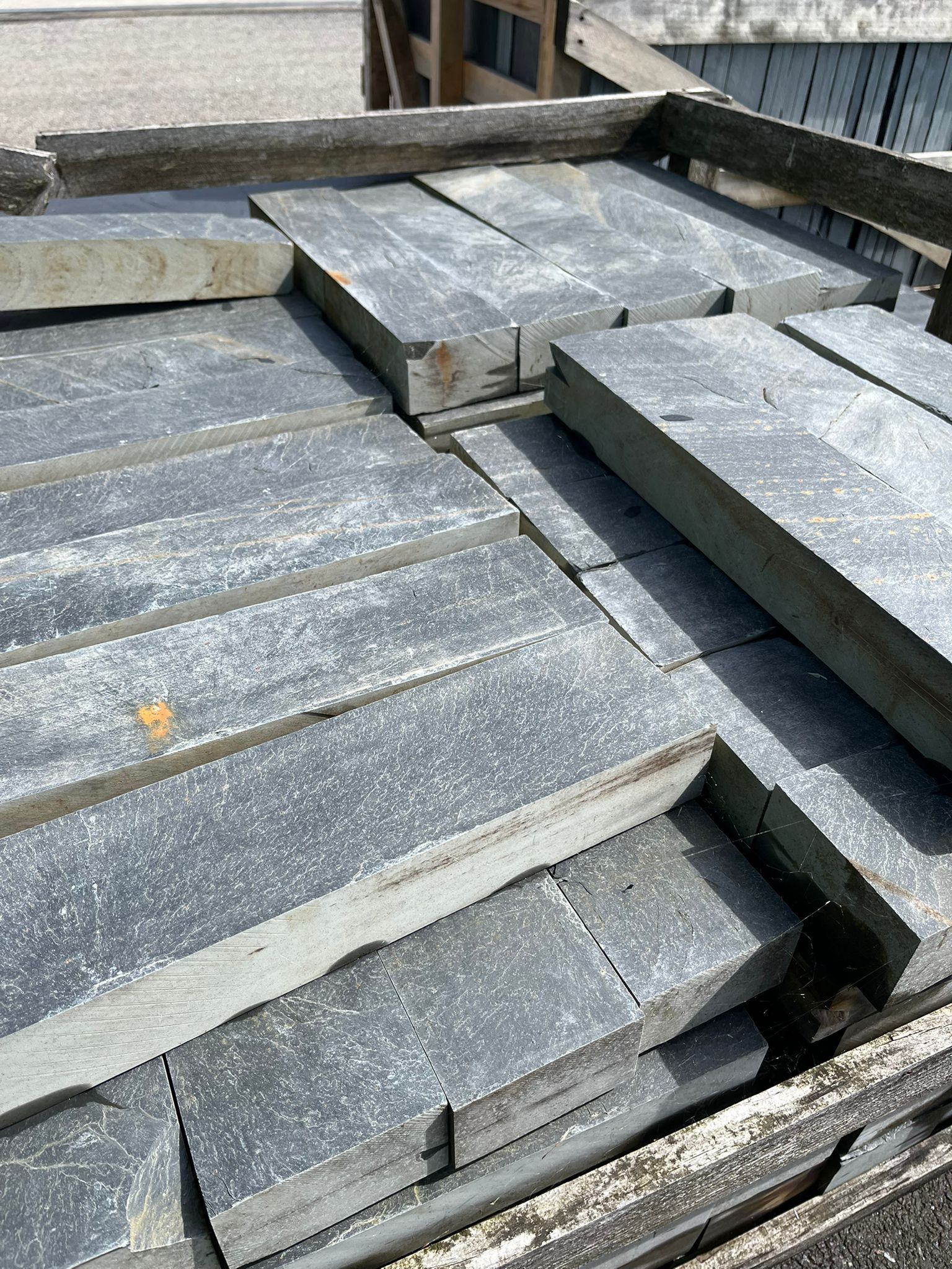 Dalles de pierre sombre empilées sur des palettes dans une cour de gravier, avec une longue bordure noire au premier plan.