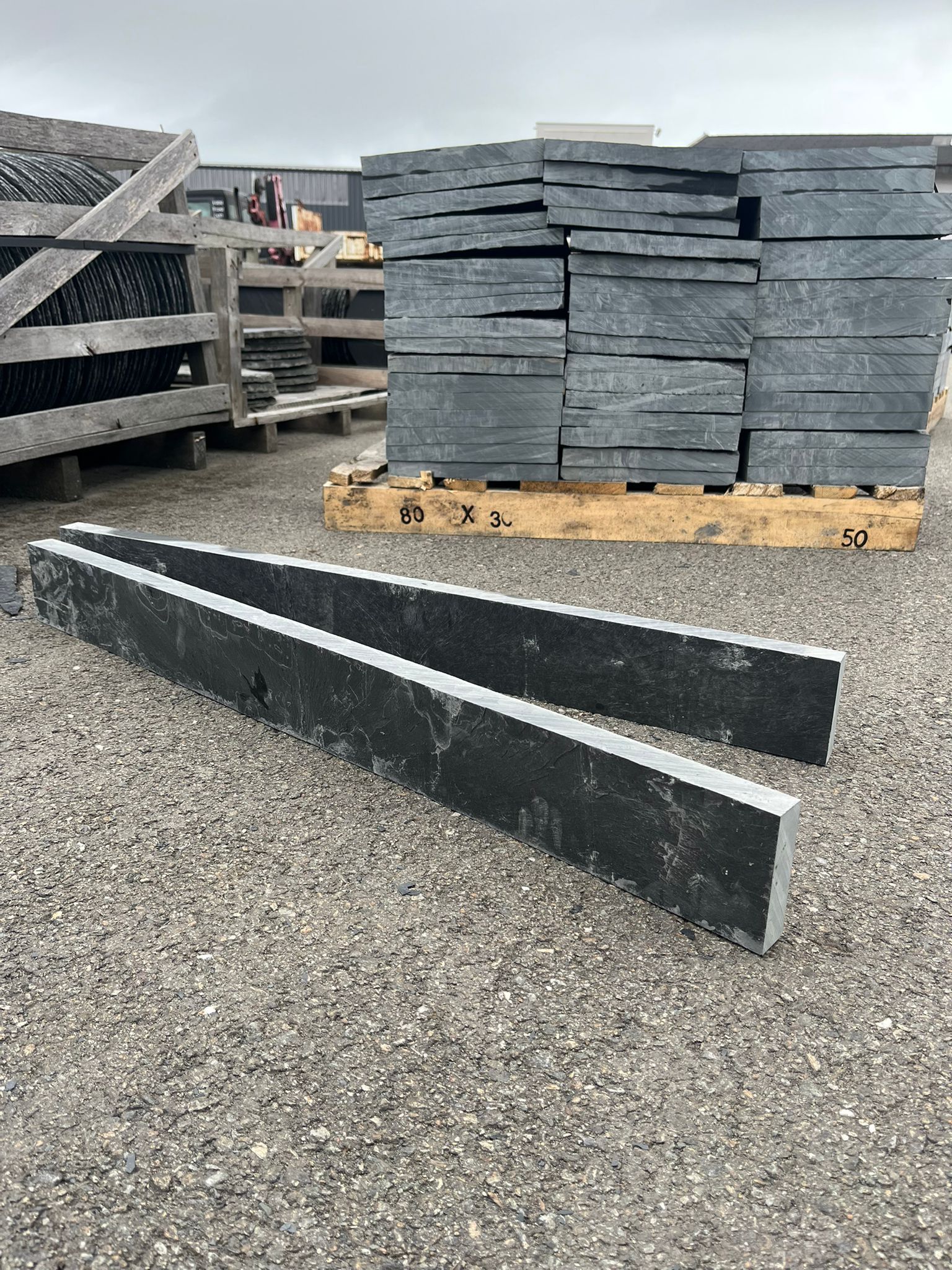 Dalles de pierre sombre empilées sur des palettes dans une cour de gravier, avec une longue bordure noire au premier plan.