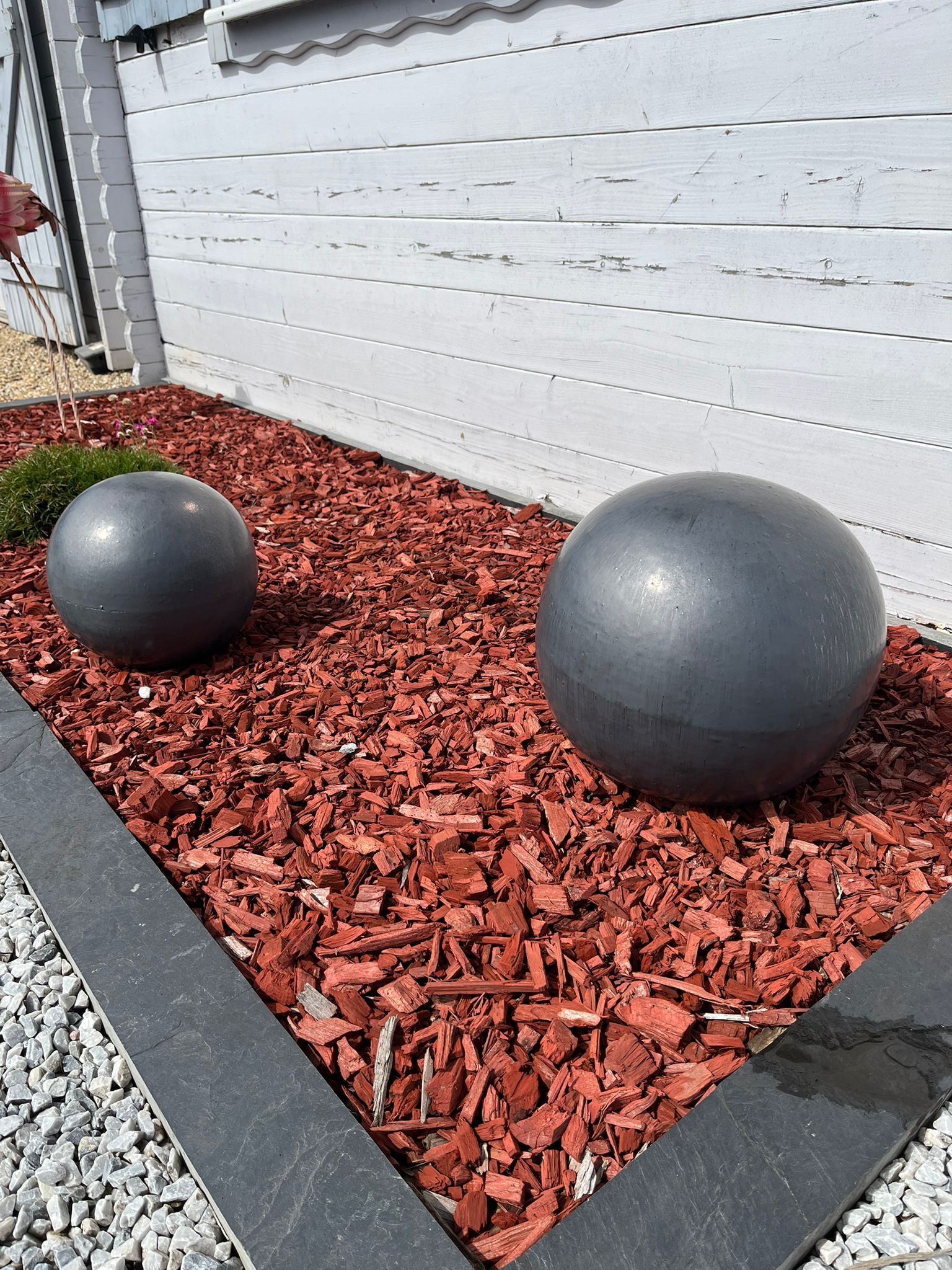 Deux sphères décoratives grises sur du paillis rouge, près d'un mur blanc et d'une bordure de pierres noires.